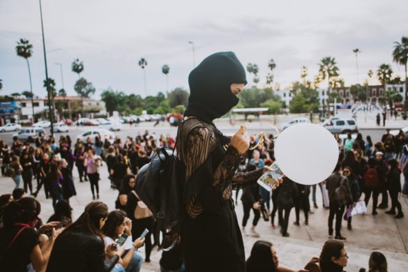 Persona con sudadera con capucha negra y mangas de encaje sosteniendo un gran globo blanco entre una multitud de personas en un entorno urbano.