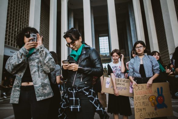 Grupo de mujeres en una protesta, algunas con carteles, una tomando una foto y otra revisando su teléfono.