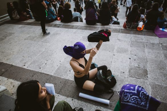 Una mujer vestida de negro y un pañuelo morado se sienta en unas escaleras, usando un teléfono inteligente, rodeada de otras personas sentadas en una reunión al aire libre.