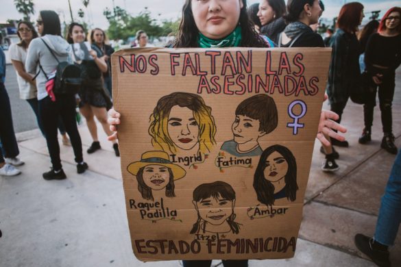 Un manifestante sostiene un cartel que representa a cinco mujeres con el texto "nos faltan las asesinadas" y "el estado feminicida" en una manifestación.