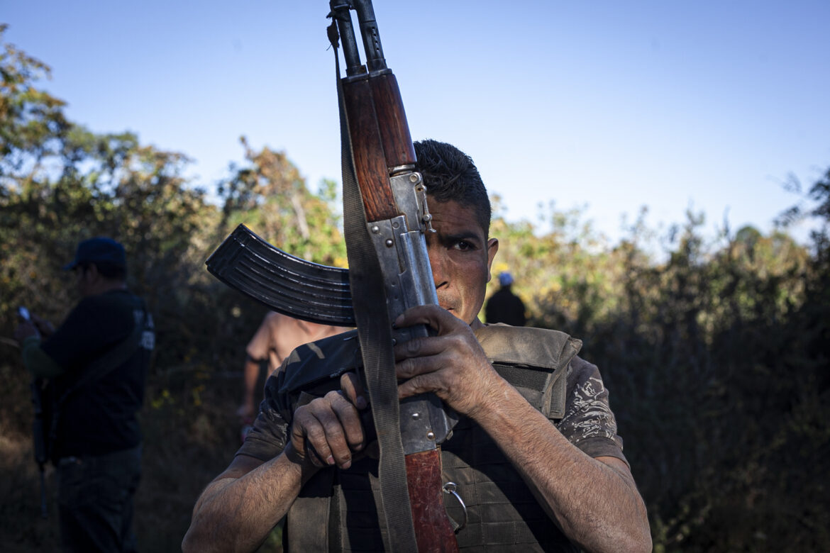 Un hombre con chaleco antibalas sostiene un rifle en una zona arbolada al aire libre, con otras personas visibles al fondo.